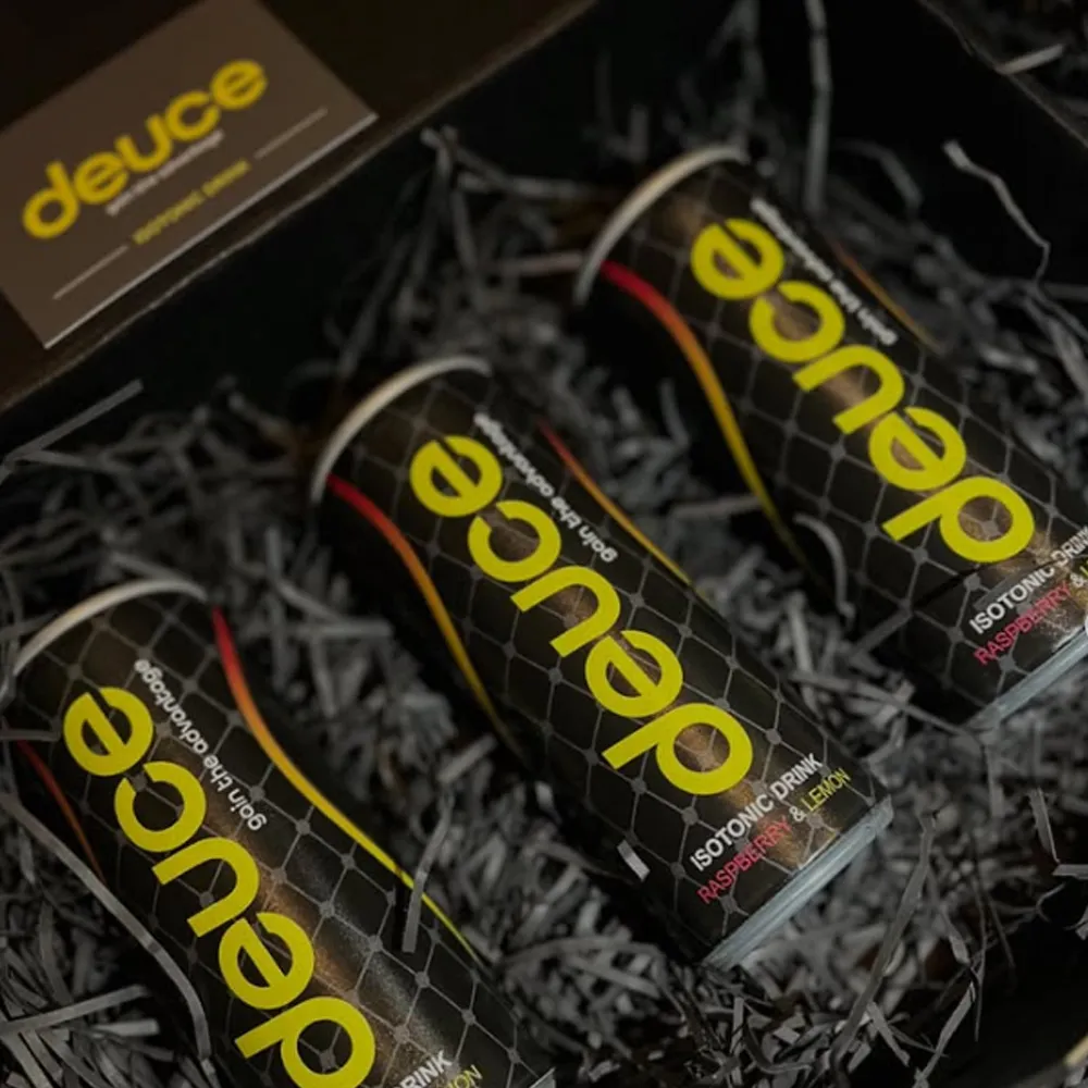 Three cans of Deuce isotonic drink in a black box filled with black shred paper. The cans are labeled raspberry and lemon.